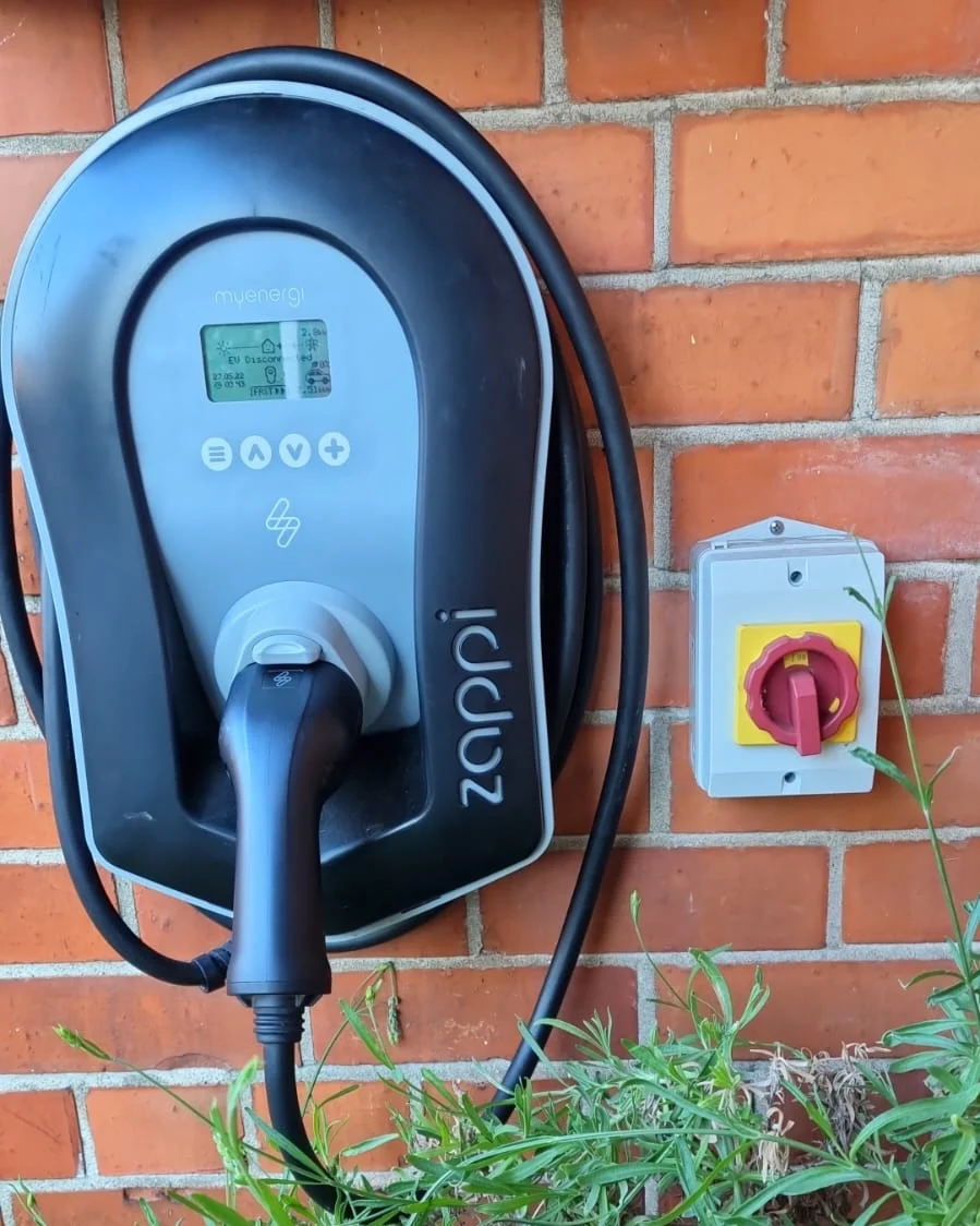 A wall-mounted EV charge point on a brick wall with its cable connected, and a red rotary isolator switch mounted alongside