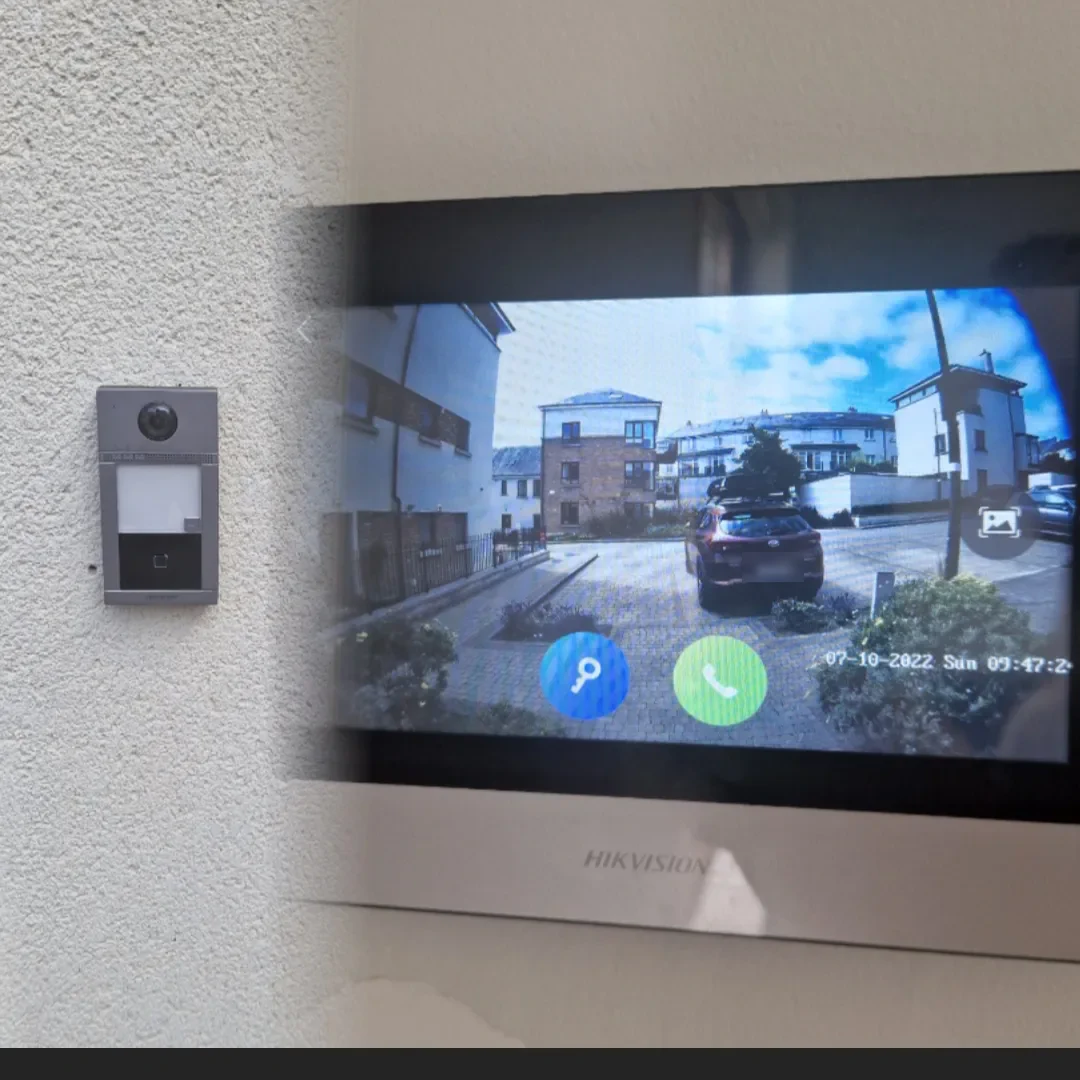 A Hikvision door entry station mounted beside an indoor monitor showing the live camera view of the approach to the door